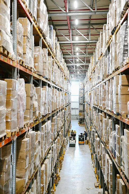 High angle photo of warehouse walkway, either side flanked by towering shelves of boxes. Image represents supply chain warehousing activity.